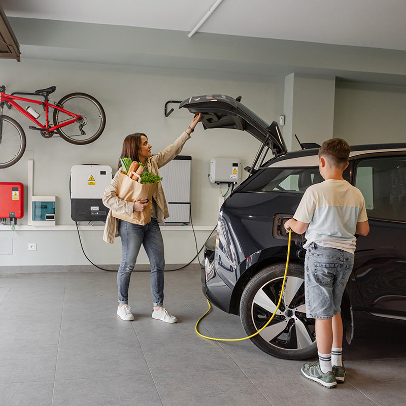 Son plugs EV charger from charging station to electric vehicle in home while mother takes groceries from the car